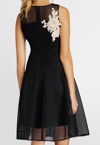 Femme portant une robe en mesh noir sans manches avec une jupe arrivant au genou et des appliqués floraux blancs dans le haut du dos.