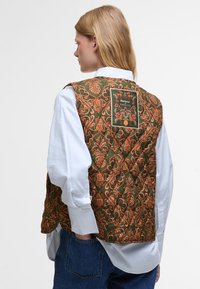 Quilted vest featuring an olive green base with an orange floral pattern, a square label, and white shirt underneath.