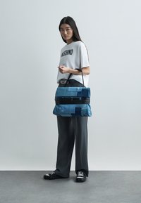 Τσάντα ώμου από τζιν με σχέδιο πατς-γουόρκ σε διάφορες αποχρώσεις του μπλε, με μαύρο φερμουάρ και διπλές λαβές για μεταφορά.