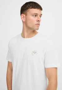 Tricou alb din bumbac, cu mâneci scurte, având un mic logo de atom bej brodat în zona stângă a pieptului. Design simplu și clasic.