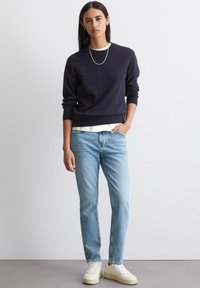 Marineblauwe trui, ronde hals, geribbelde manchetten, over een licht shirt gedragen. Lichtblauwe jeans, rechte pasvorm, en witte sneakers met een stevige zool.