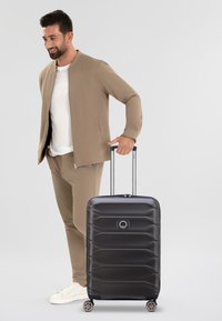 Maleta rígida negra con textura ondulada, manija retráctil plateada y cuatro ruedas. Acompañada por una persona vestida con un atuendo beige.