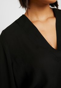 Black V-neck top with a smooth texture, featuring wide sleeves and a simple design. The neckline forms a subtle V-shape.