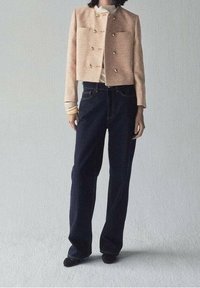 Veste courte texturée beige avec des boutons dorés associée à un jean droit bleu foncé. Le mannequin porte un haut clair en dessous.