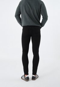 Person som står med händerna i fickorna iklädd en mörkgrå tröja, svarta skinny jeans och svarta sneakers med vita ränder framför en enkel bakgrund.