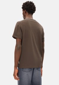 Mann mit kurzen, lockigen Haaren, der ein braunes T-Shirt mit kurzen Ärmeln und blaue Jeans trägt, von hinten vor einem einfarbigen Hintergrund gesehen.