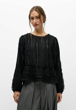 Modèle portant un chemisier noir texturé à manches longues et un pantalon gris plissé, sur un fond blanc uni.