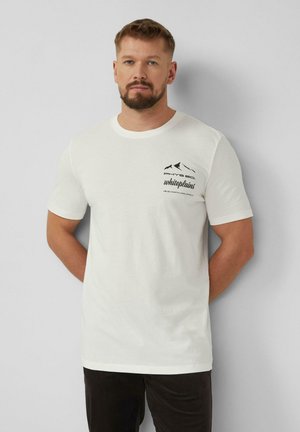 Mann mit kurzen Haaren und Bart, trägt ein weißes T-Shirt mit Berg-Logo und Schriftzug „PHYS ED Whiteplains“, steht vor einer hellgrauen Wand.