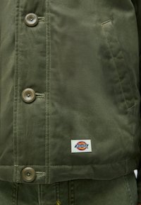 Olivno zelena jakna z gumbi, ki ima žep in nalepko Dickies. Gladka tekstura in klasična zasnova ovratnika.