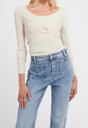 Kvinde iført en beige langærmet top med en rund ringdetalje ved brystet og højtaljede blå jeans med synlige knapper og forlommer.