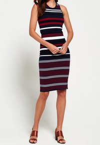 Armoton tummansininen mekko, jossa on punaisia ja valkoisia vaakasuoria raitoja. Liukuväriinen, polvipituinen siluetti, valmistettu joustavasta kankaasta. Ruskeat sandaalit täydentävät asun.