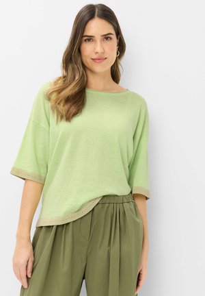 Kvinde med langt brunt hår iført en lysegrøn kortærmet top og løse olivengrønne bukser mod en ensfarvet hvid baggrund.