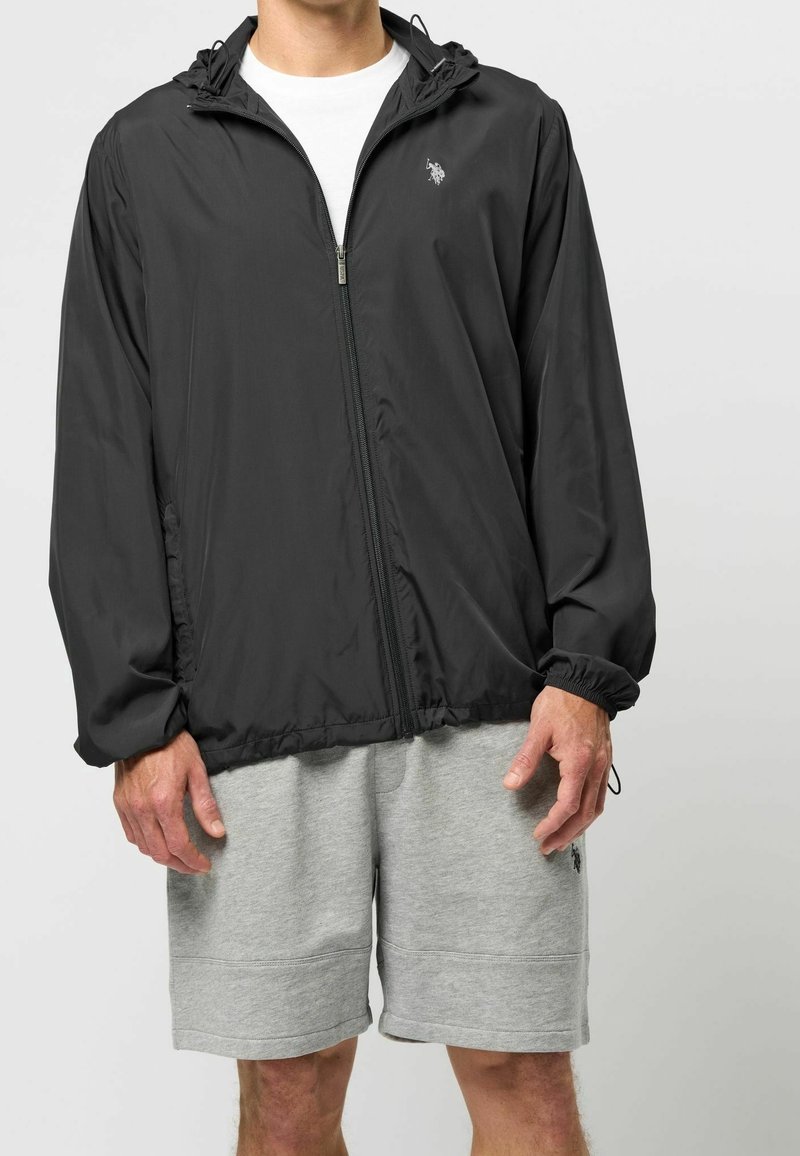 Svart lättvikts zip-up jacka med huva, elastiska muddar och en diskret logo. I kombination med grå bomullsshorts. Slät textur, avslappnad passform.