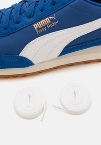 Zapatilla azul con acentos blancos, fabricada con materiales de ante y sintéticos. Incluye dos cordones blancos con puntas de plástico al lado de la zapatilla.