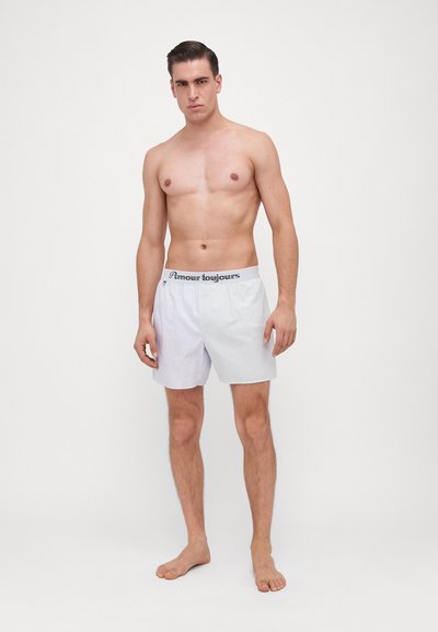 Boxer shorts in light gray with thin vertical white stripes. Elastic waistband with "Amour Toujours" in black. Model stands barefoot, shirtless.