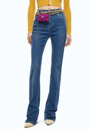 Donna che indossa jeans a zampa a vita alta blu, maglione corto giallo con polsini in pizzo, scarpe beige con punta e cintura a catena dorata con una piccola pochette viola.