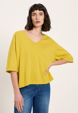 Woman with shoulder-length dark hair wearing a loose yellow V-neck top and blue jeans, posing with one hand on hip against plain background.