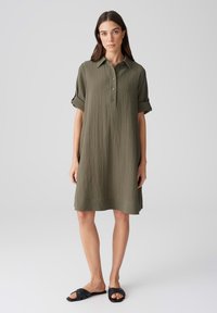 Abito camicia verde oliva, lunghezza al ginocchio, con tessuto strutturato, chiusura a bottoni e maniche arrotolate. Abbinato a sandali neri con infradito.
