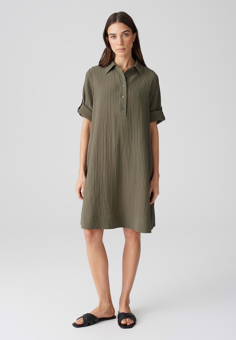 Abito camicia verde oliva, lunghezza al ginocchio, con tessuto strutturato, chiusura a bottoni e maniche arrotolate. Abbinato a sandali neri con infradito.