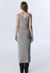 Kobieta widziana z tyłu, ubrana w szary, bezrękawnikowy sweter z falbaniastym dekoltem oraz czarne kozaki do kolan na białym tle.