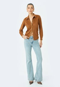 Camisa marrón de botones con mangas largas, combinada con jeans de campana azul claros y tacones de leopardo. Textura suave, silueta clásica.