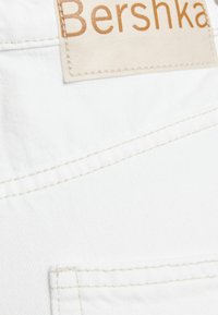 Bílá denimo tkanina s viditelným šitím; obsahuje béžovou nášivku s embosovaným nápisem "Bershka" ve zlaté barvě. Čisté linie a minimalistické designové prvky.