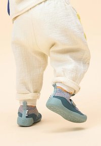 Blaue und grüne Kinderschuhe mit strukturierten Sohlen, begleitet von mehrfarbigen, gestrickten Knöchelsocken und cremefarbenen, hochgekrempelten Hosen.