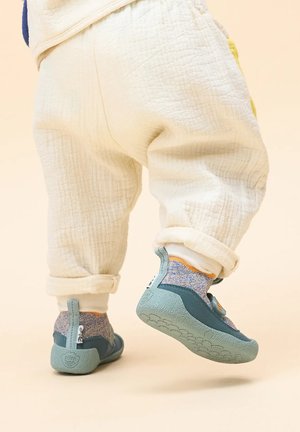 Blaue und grüne Kinderschuhe mit strukturierten Sohlen, begleitet von mehrfarbigen, gestrickten Knöchelsocken und cremefarbenen, hochgekrempelten Hosen.