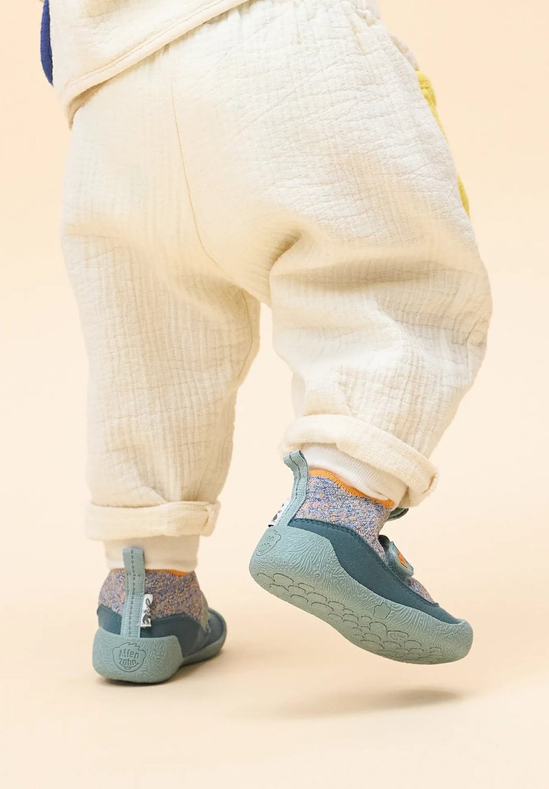 Blaue und grüne Kinderschuhe mit strukturierten Sohlen, begleitet von mehrfarbigen, gestrickten Knöchelsocken und cremefarbenen, hochgekrempelten Hosen.