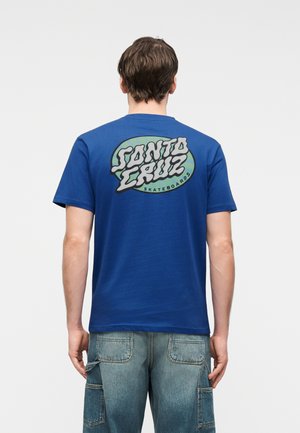 Mand iført en blå Santa Cruz Skateboards t-shirt og blå jeans, set bagfra mod en ensfarvet baggrund.