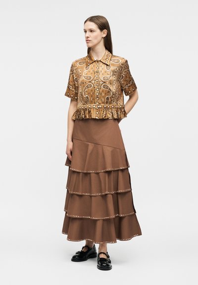 Γυναίκα που φοράει καφέ πουκάμισο με κοντά μανίκια και πασλεδάτο μοτίβο, με βολάν στο τελείωμα, και καφέ επιπέδων φούστα έως το μέσο της γάμπας με διακοσμητικές καρφίτσες στις άκρες.