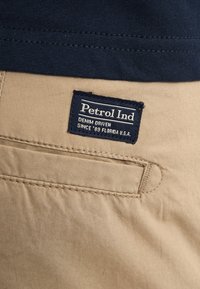 Material bej cu o textură netedă, având o etichetă albastru închis pe care scrie „Petrol Ind”, detaliind „DENIM DRIVEN” și „SINCE '89 FLORIDA U.S.A.”.