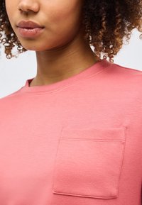 Κοντινό πλάνο ενός ατόμου με σγουρά μαλλιά που φοράει ένα ροζ πουκάμισο με μια μικρή τετράγωνη τσέπη στο στήθος στην αριστερή πλευρά.