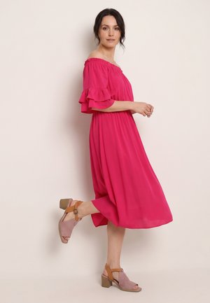 Woman wearing a bright pink off-shoulder midi dress with ruffled sleeves and beige block heel sandals standing against a plain background.