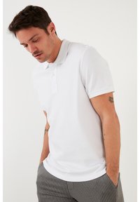 Camisa polo blanca de canalé con cuello, que cuenta con una tapeta de tres botones. Combinada con pantalones grises de rayas, que muestran un ajuste casual.