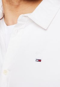 Ingrandimento di una persona che indossa una camicia bianca con bottoni, con un piccolo logo rettangolare rosso, bianco e blu sul petto.