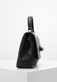 Sac à main en cuir noir au design structuré, surface texturée, quincaillerie en argent et un charm décoratif amovible. Vue de côté.