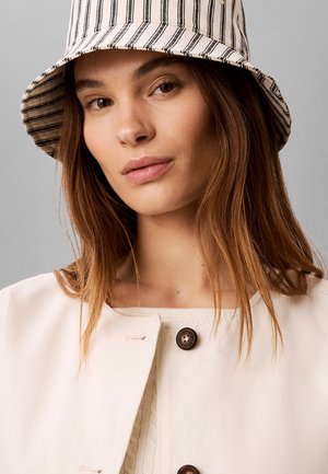 Giovane donna con lunghi capelli castani che indossa un cappello a secchiello a righe e una giacca crema con bottoni davanti a uno sfondo grigio.