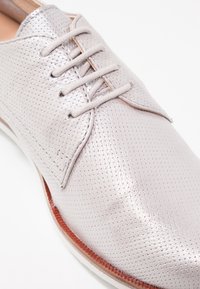 Chaussure à lacets en cuir gris clair perforé avec bordure marron et semelle blanche, vue en gros plan.