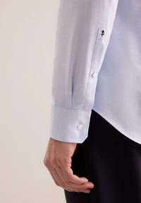 Camicia a maniche lunghe di colore azzurro chiaro con tessuto testurizzato, polsini con bottoni e un piccolo fiore nero ricamato sul polsino.