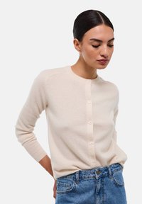 Cardigan en maille crème avec un col rond, des manches longues et une fermeture à six boutons sur le devant. Texture douce et matériau léger. Porté par-dessus un jean.