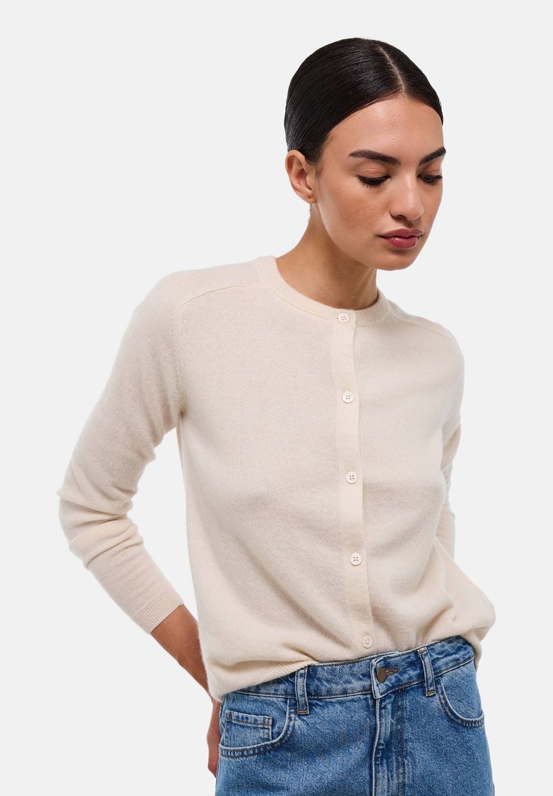 Cardigan en maille crème avec un col rond, des manches longues et une fermeture à six boutons sur le devant. Texture douce et matériau léger. Porté par-dessus un jean.