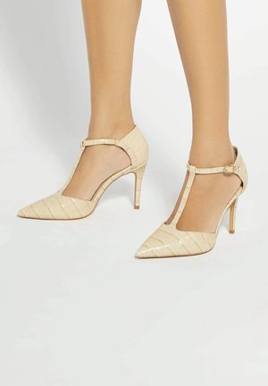 Piernas con zapatos de tacón alto color beige con textura de cocodrilo, punta puntiaguda y tiras en T con hebillas en el tobillo, sobre fondo blanco.