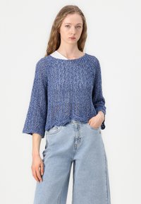 Pull en laine bleu avec une coupe ample, des manches larges et un ourlet festonné, porté par-dessus un haut blanc, associé à un pantalon en denim bleu clair à jambes larges.
