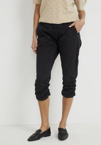 Svarta korta byxor med rynkade muddar och sidofickor. Tyget verkar lätt och har en slät struktur. Kombineras med svarta loafers.