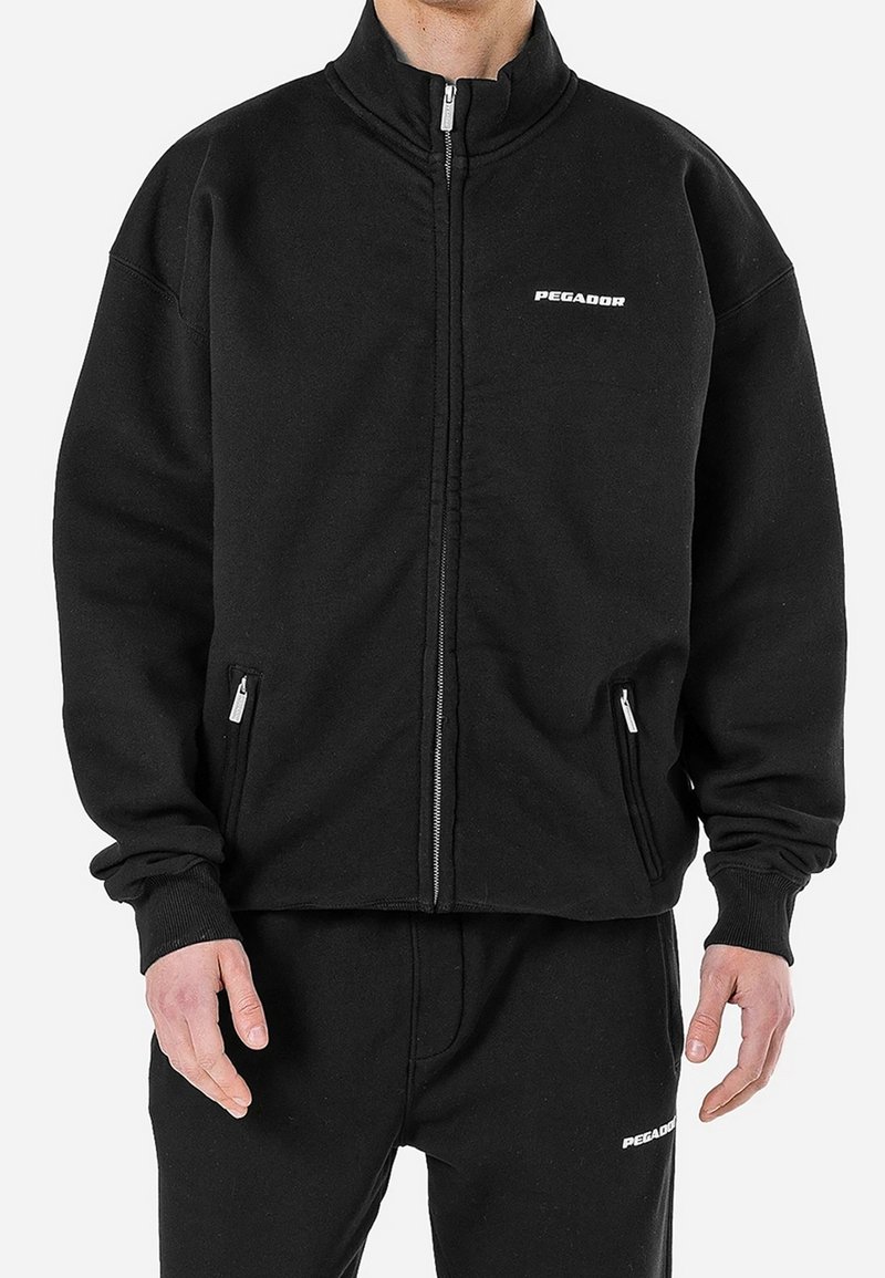 Homme portant une veste noire zippée et un pantalon avec des logos blancs "PEGADOR" sur la poitrine et la cuisse.