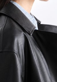 Giubbotto di pelle nera con colletto a punta, texture liscia e design drappeggiato, indossato sopra una camicia azzurro chiaro con colletto visibile.