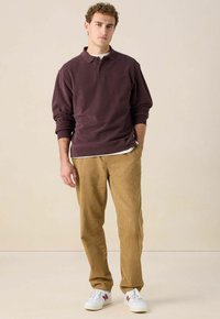 Junger Mann in einem weinroten Langarm-Polo, beigen Cordhosen und weißen Sneakers, der vor einem schlichten beigen Hintergrund steht.