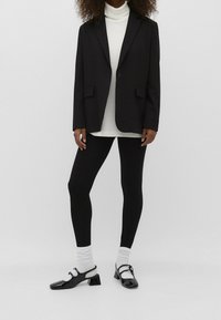 Blazer nero sopra un dolcevita bianco, abbinato a leggins neri e scarpe in vernice nere con punta quadrata e tacco a blocco.