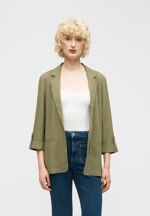 Personne blonde portant un blazer ouvert vert olive sur un haut blanc et un jean bleu, debout devant un fond uni gris clair.